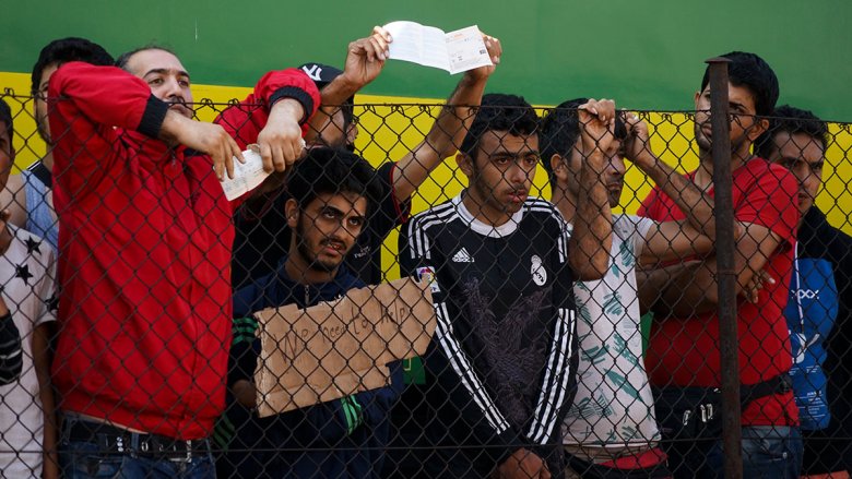 Male refugees hold up signs behind a fence
