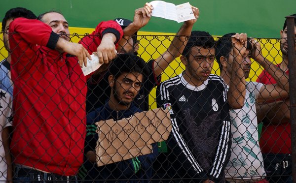 Male refugees hold up help signs behind a fence
